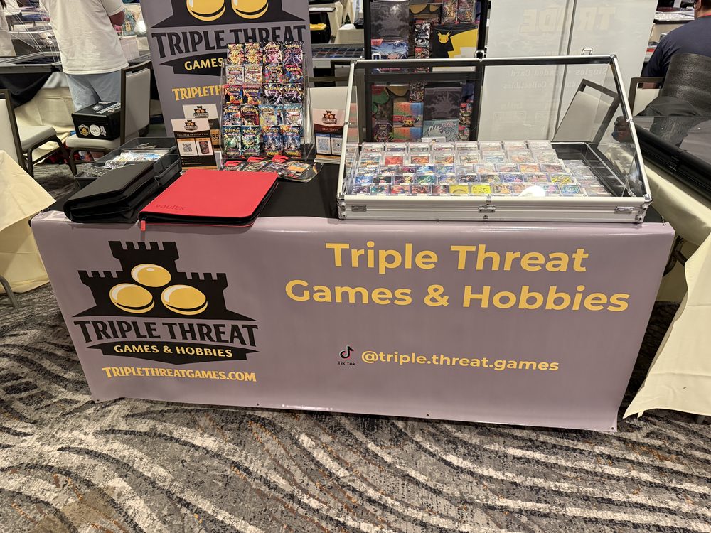 Triple Threat Games & Hobbies booth display at a card show, with graded cards and sealed product on the table.